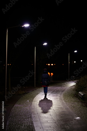 Postać kobiety tyłem idącej alejką w parku miejskim oświetlona latarniami nocą