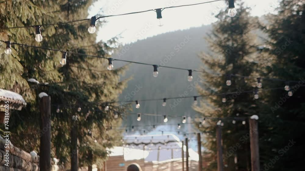 A beautiful young girl tourist walks through a snowy village high in the mountains. Lanterns and light bulbs on the street against the backdrop of forests and houses. Fresh air on holiday for winter