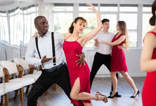 Emotional elegant adult African American performing kizomba paired with attractive woman in red, demonstrating dance characteristic sensual and connected movements in studio class setting..