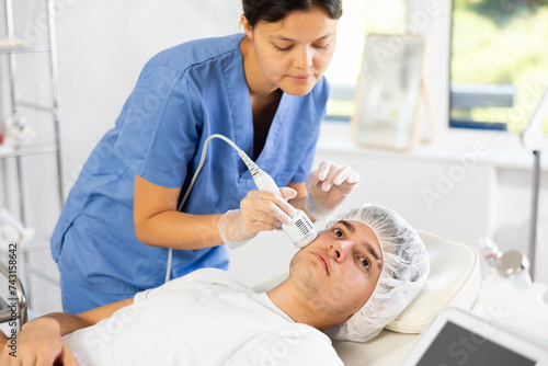 Young woman cosmetologist performs facial whitening procedure with apparatus young male patient
