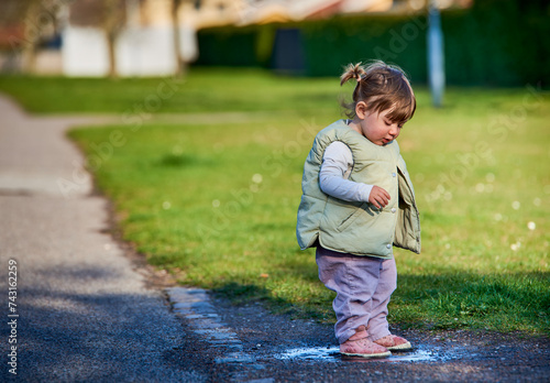 Rascal little female child with braids jumping in a muddy puddle on a sunny spring day. Scoundrel 2 year old girl hopping in a small water plash by the grass. Kids exploration and freedom concept