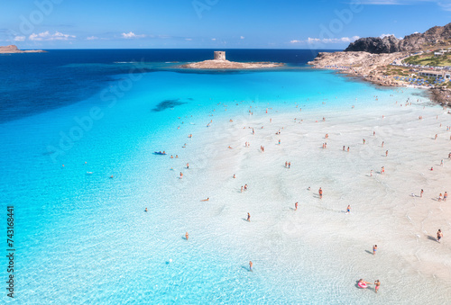 Aerial view of  La Pelosa beach at sunny summer day. Stintino, Sardinia island, Italy. Top view of white sandy beach, swimming people, clear blue sea, old tower and sky with clouds. Tropical seascape