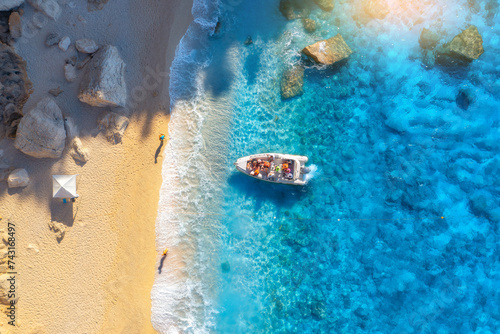 Aerial view of boat, sandy beach and blue sea with waves at sunset. Summer in Sardinia, Italy. Top drone view of motorboat, shore, stones in clear water. Colorful tropical landscape. Travel. Seaside