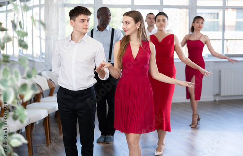 Elegant couple, smiling young guy and stylish woman in red dress enjoying slow foxtrot in dance studio. Amateur ballroom dancing concept..