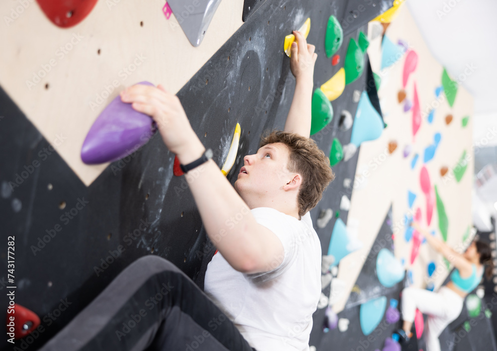Fototapeta premium Teenage boy is mastering climbing on training wall in gym, side view. Teen guy holds on tightly to ledges and strives for top of bouldering route