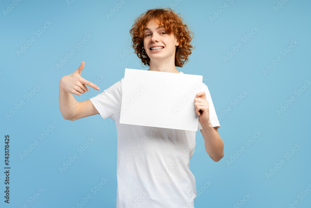 Naklejka premium Good looking redheaded male teen with braces, pointing at a banner, isolated on blue background
