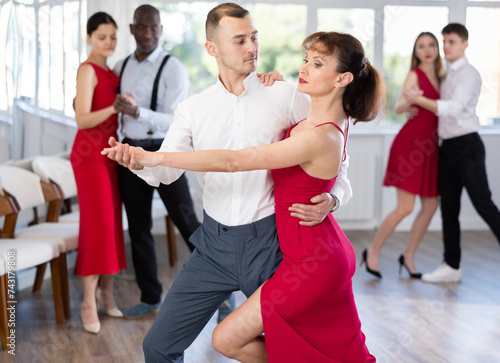 Elegant couple, attractive woman in red dress and stylish man, performing expressive tango in bright, modern dance studio during group class..
