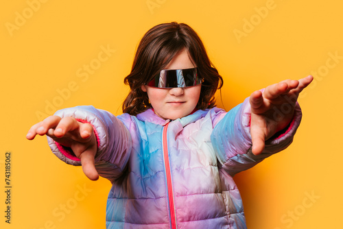 Confident girl posing with futuristic glasses and extending her hands forward, wearing a colorful pastel jacket against an orange background