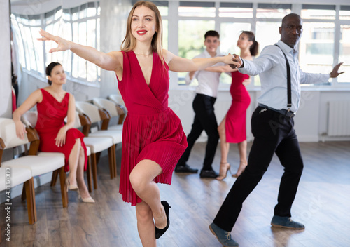 Positive adult woman in vibrant red dress enjoying active dances with black male partner in modern dance studio, practicing playful jitterbug moves..