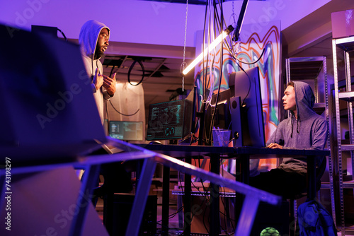 Two diverse hackers working together to hack computer system in abandoned warehouse. Asian and african american internet criminals discussing malicious software code in dark workspace