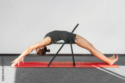 Photos Yoga practitioner in sportswear, chair used as support to perform prasarita parsvakonasana pose