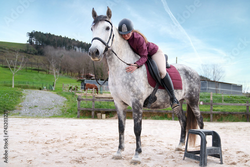 Focused young rider mounting a speckled white horse in an open sandy arena before starting equestrian practice © Koldo_Studio