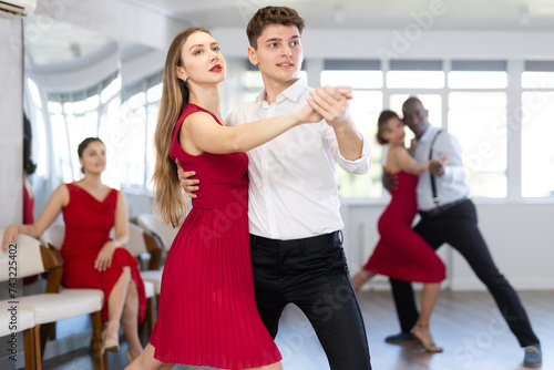 Positive modern young guy in formal attire and woman wearing elegant red dress gracefully waltzing during group slow ballroom dancing class in choreography studio