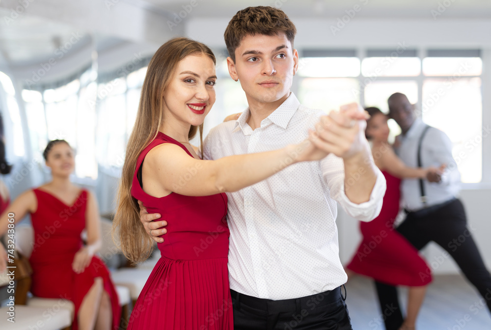 Pretty young woman dancing waltz with partner at group of multinational people in modern ballroom dancing class