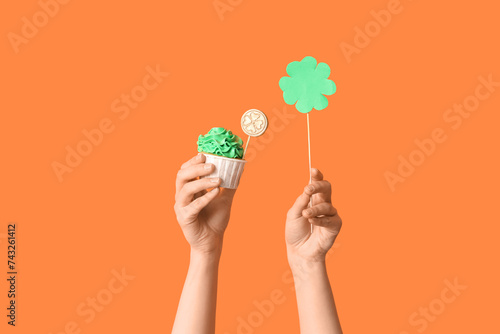 Female hands with tasty cupcake and paper clover for St. Patrick's Day on orange background