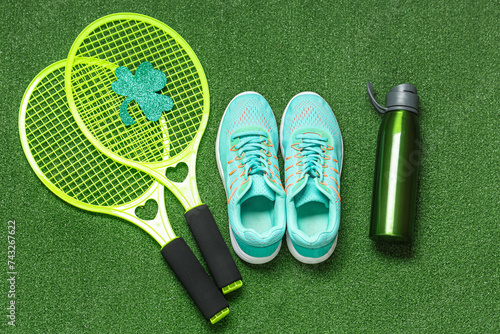 Sneakers, tennis rackets, water bottle and clover on green grass background. St. Patrick's Day celebration
