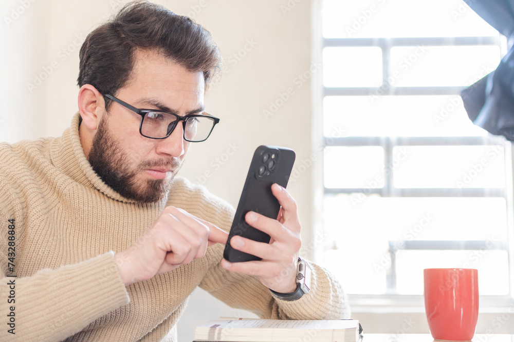 Fototapeta premium Joven apuesto con gafas revisando su celular desde la comodidad de su hogar. 
