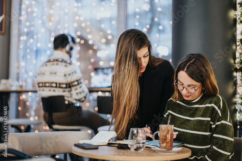 Two friends engaged in a conversation at a warmly lit cafe. The bokeh lights create a holiday mood while one woman points to a tablet screen.