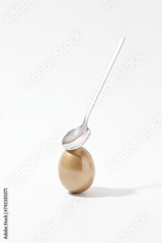 Minimal scene of a golden Easter egg and a spoon decorated against white background. Easter commemorates the resurrection of Jesus Christ, three days after he was crucified