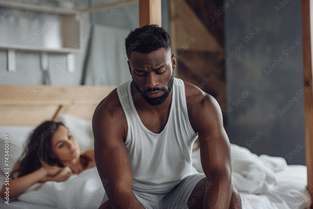 handsome black man sitting on the edge of his bed worried expression, in the background a woman looking at him. relationship problems handsome black man sitting on the edge of his bed worried expression, in the background a woman looking at him. relationship problems