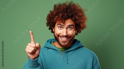 A cheerful smiling curly-haired man with stubble, wearing casual clothes, points with his index finger at an empty space for your slogan, advertising on a green background.