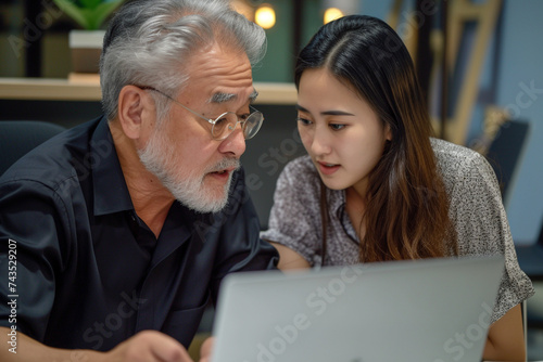 Mature Latin manager mentor talking to young Asian female coworker showing online project results at meeting. Two happy diverse professional executives team working in office using pc laptop