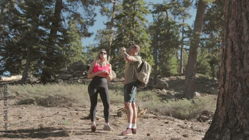 A male hiker in green shorts and a backpack drinking water on a break on a forested trail on a bright day with his girl friend. Slow Motion, 4K RAW. A male hiker in green shorts and a backpack drinking water on a break on a forested trail on a bright day with his girl friend. Slow Motion, 4K RAW.