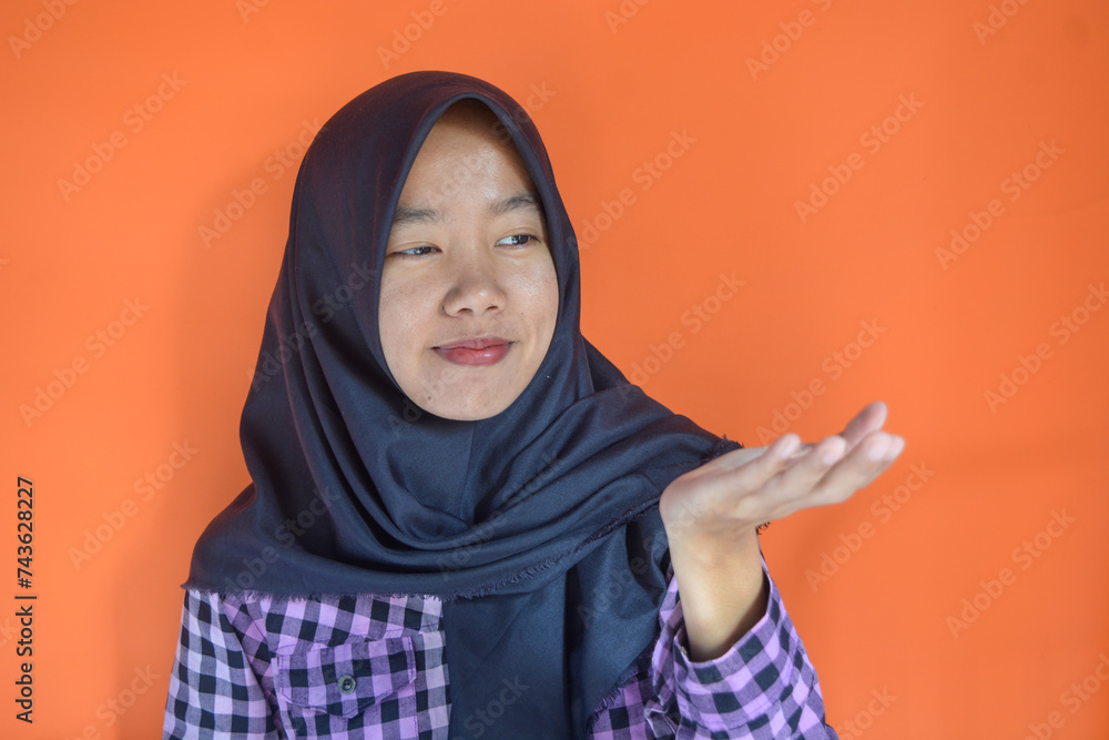 An Asian Muslim woman is wearing a purple shirt with a black hijab. Is pointing in a direction with an orange background An Asian Muslim woman is wearing a purple shirt with a black hijab. Is pointing in a direction with an orange background
