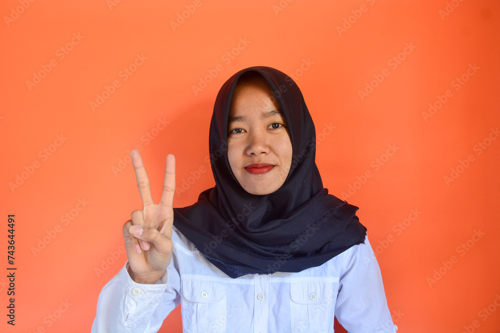 Asian Muslim woman close up raising two fingers with smiling face on blurred orange color textured background with copy space Asian Muslim woman close up raising two fingers with smiling face on blurred orange color textured background with copy space
