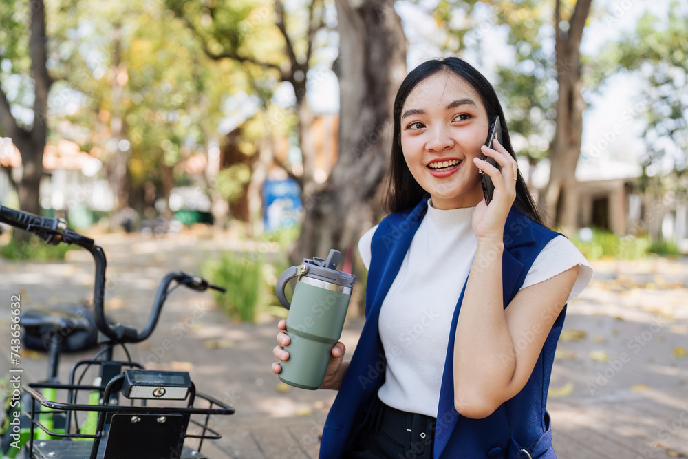 Fototapeta premium Smiling asian businesswoman hold reusable eco-friendly ecological cup and using mobile while sitting at park