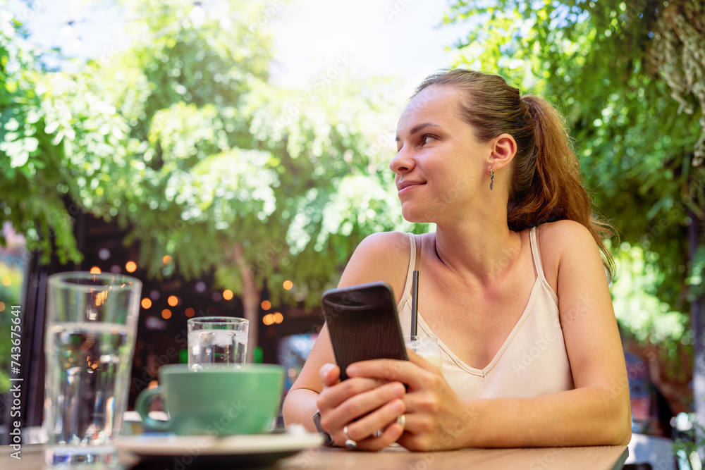 Brunette woman sits at the table at cafe holds smartphone in hands and smiling. Brunette woman sits at the table at cafe holds smartphone in hands and smiling.