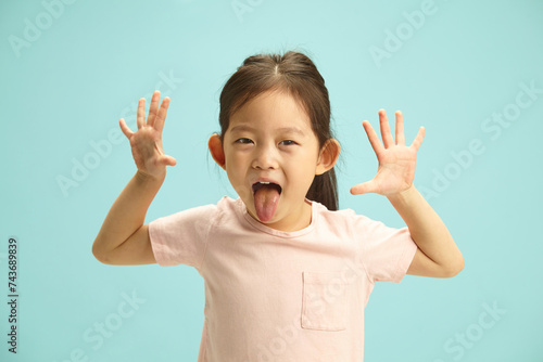 Playful and Lovely Asian Ethnicity Child Girl cheerfully showing opening hands up, sticking out tongue, expresses joyful mood standing against blue isolated. Concept of children happy.
