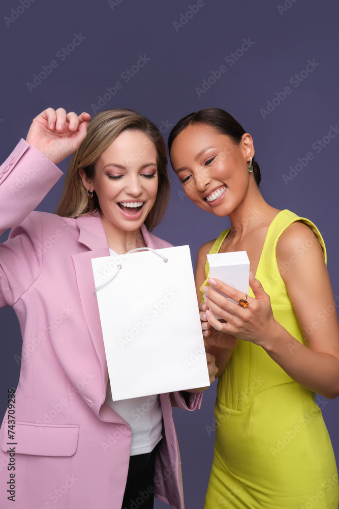 two beautiful Asian and European girls open a gift in a white box in beautiful jewelry on a blue background. cute girls are dressed in stylish dresses. pleasantly surprised two beautiful Asian and European girls open a gift in a white box in beautiful jewelry on a blue background. cute girls are dressed in stylish dresses. pleasantly surprised