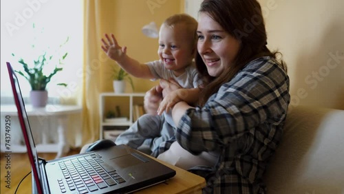 Happy smiling Caucasian woman with cute baby waving hands, chatting online on laptop sitting on couch. The concept of online communication, happy family, digital lifestyle. Modern living room