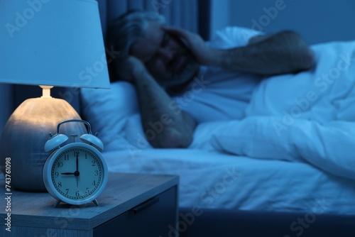 Mature man suffering from headache in bed at night, focus on alarm clock photo
