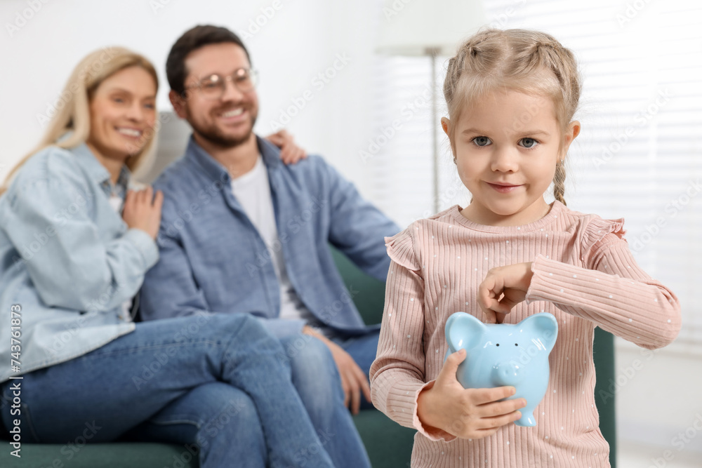 Family budget. Little girl putting coin into piggy bank while her parents watching indoors, selective focus Family budget. Little girl putting coin into piggy bank while her parents watching indoors, selective focus