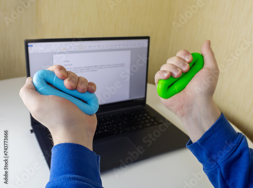 Hand gripper. Man squeezes rubber expander while working on his laptop. Concept of combining useful activity with necessary work. Training hand and arm muscles. Multi-colored mini training apparatus photo