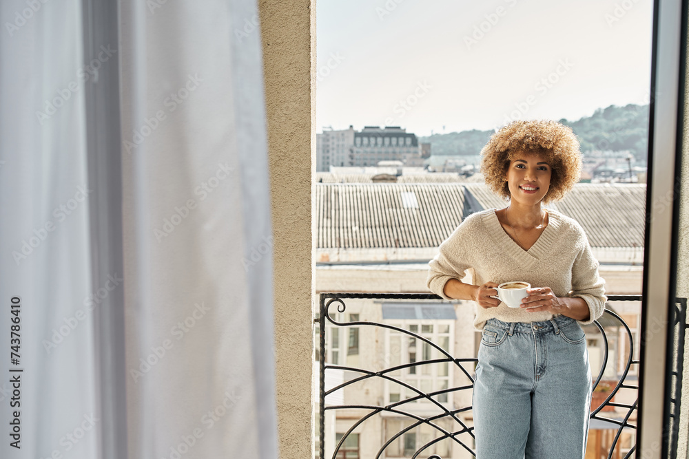 Fototapeta premium smiling curly african american woman standing with cup of coffee on hotel balcony, city view