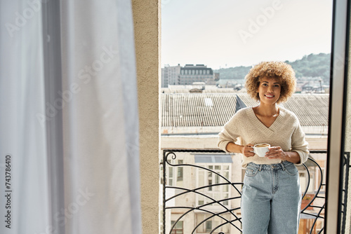 smiling curly african american woman standing with cup of coffee on hotel balcony, city view © LIGHTFIELD STUDIOS