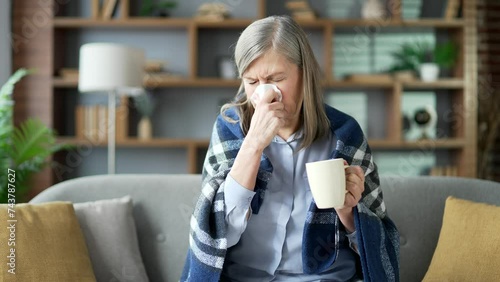 Sick senior female wrapped in a blanket suffers from a runny nose while sitting on sofa in living room at home. Upset elderly woman wipes her nose with a handkerchief and drinks a hot drink from a cup photo