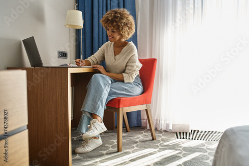 happy and curly african american woman remote working in hotel room, using laptop and taking notes photo