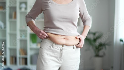 Midsection of woman hands holding her belly fat. Overweight female with excess weight touching and squeezing cellulite on her big waist standing at home. Concept of obesity, overweight and overeating photo