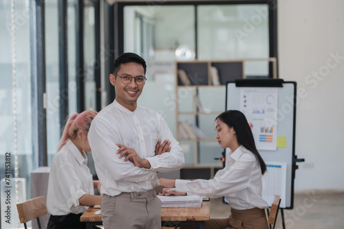 Smiling young Asian businessman standing in the office room with coworkers in the background.
