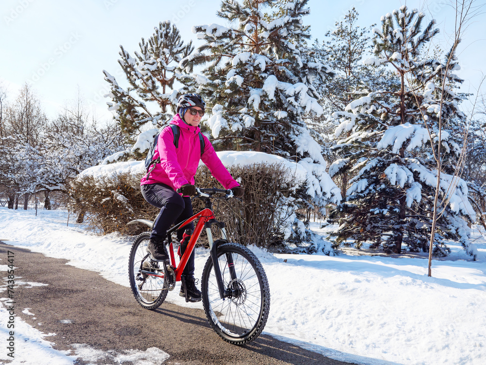 A girl in a pink jacket rides a bicycle along a path in the park after a snowfall. Winter cycling adventure. Active lifestyle