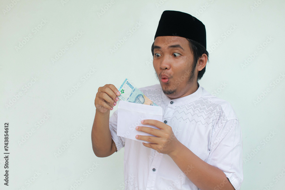 A young Asian muslim man holding an envelope and fifty thousand rupiah in cash with an expression of surprise and happiness. THR (Tunjangan Hari Raya) or Religious Holiday Allowance Concept. A young Asian muslim man holding an envelope and fifty thousand rupiah in cash with an expression of surprise and happiness. THR (Tunjangan Hari Raya) or Religious Holiday Allowance Concept.