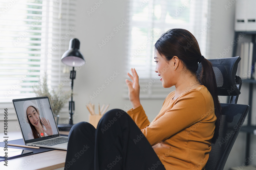 A woman in a casual mustard top waves greeting during a friendly video call on her laptop in a home office.