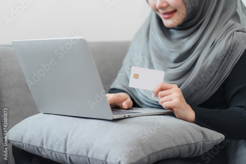 Confident Muslim woman in a hijab showing a credit card mockup while using a laptop, preparing for an online purchase.