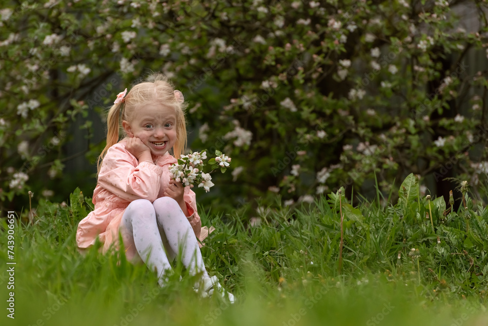 Happy smiling little blonde girl with two ponytails holds blooming flowers and sits on grass. Spring day