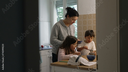 Candid domestic lifestyle scene of mother standing in kitchen with her two children preparing meal together. Small brother and sister interacting with parent while she pours honey into bowl