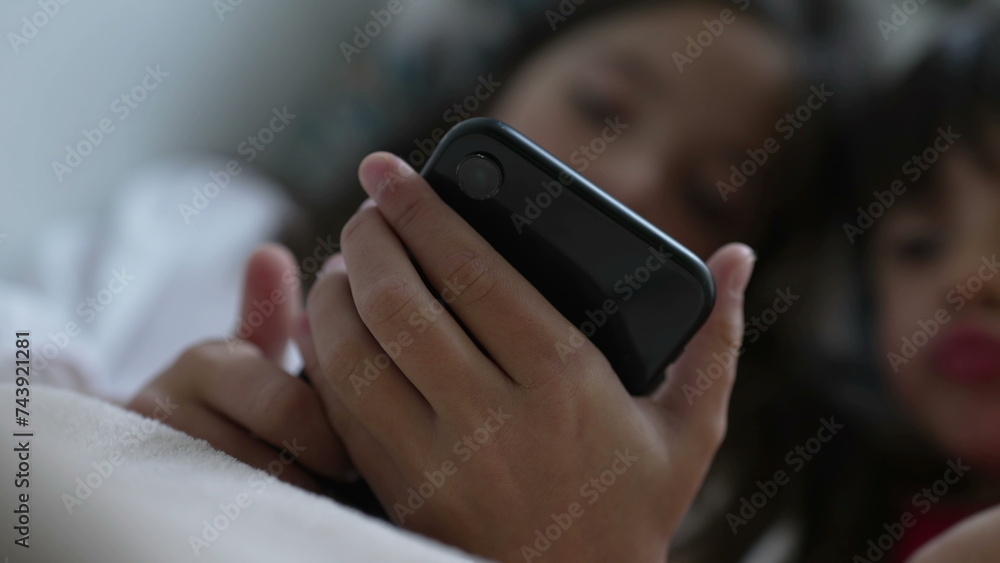Child close-up hand holding cellphone device absorbed by media entertainment online, blurred kids in background staring at phone screen - obrazy, fototapety, plakaty 